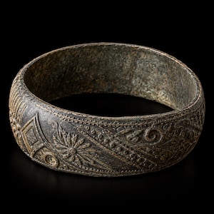 Może przedstawiać: Ciemna, spatynowana metalowa bransoleta typu bangle o teksturowanej powierzchni z misternymi, tłoczonymi wzorami geometrycznymi, w tym kształtami rombów, motywami kwiatowymi i kropkowanymi liniami, które biegną wokół całego obwodu obręczy.