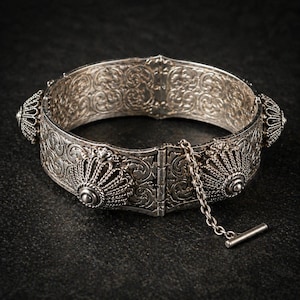 Könnte beinhalten: Ein silberfarbenes, aufklappbares Armband mit aufwendigen Rankenmustern und dekorativen Elementen. Das Armband verfügt über einen Verschluss mit Sicherheitskette und dekorativen Endstücken. Das Design weist detaillierte Muster und Texturen auf, die einen Vintage- oder Antik-Look erzeugen.