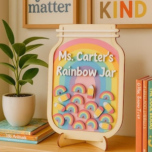 以下が含まれることがあります： 「Ms. Carter's Rainbow Jar」の文字とカラフルな虹と鉛筆が描かれた木製の虹の瓶。瓶は木製のスタンドに置かれています。「you matter」と「BE KIND」という言葉が書かれた2つの額入りのプリントが背景にあります。