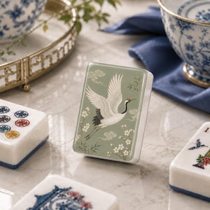 May include: A rectangular mahjong tile standing upright, featuring a sage green face with a white crane in flight, surrounded by white cherry blossoms, bamboo stalks, and stylised clouds. The tile has a smooth, glossy finish and white sides.