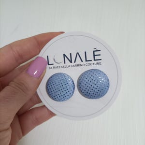 Può includere: Un paio di orecchini a bottone azzurri con un motivo a pois e un effetto scintillante. Gli orecchini sono su una carta circolare bianca con il marchio "LONALÈ" e il testo "BY RAFFAELLA CARRINO COUTURE".