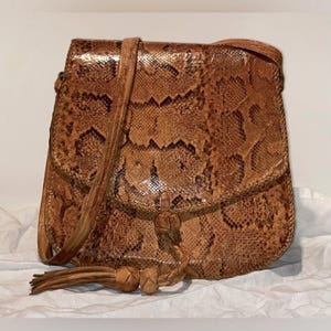 May include: A brown and tan snakeskin-effect crossbody bag with a long strap. The bag features a flap closure and a decorative tassel. The bag is made of a textured material and appears to be vintage.