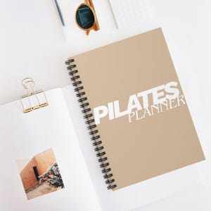 Könnte beinhalten: Ein Spiral-Pilates-Planer mit beigefarbenem Einband und weißem Text. Der Text lautet "PILATES PLANNER". Der Planer ist zu einer leeren Seite mit einem Foto geöffnet. Sonnenbrille und eine goldene Büroklammer sind ebenfalls sichtbar.
