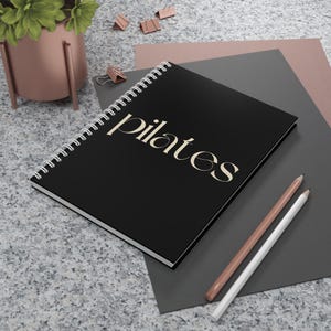 Puede incluir: Un cuaderno negro con espiral con la palabra "pilates" en escritura color crema. Dos lápices, uno blanco y otro melocotón, están junto al cuaderno. El cuaderno está sobre una superficie gris con un fondo rosa y gris.