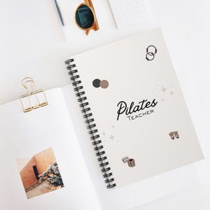 Puede incluir: Un cuaderno blanco con espiral con la frase "Pilates Teacher" en escritura negra. La portada presenta elementos decorativos y unas gafas de sol. Un clip dorado y una fotografía son visibles en la página abierta.