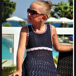 May include: A navy blue knitted baby sweater with white and red trim. The sleeveless tunic has a decorative white band across the midsection. Text on the left reads "Seamless Baby Sweater" and text on the right reads "Knitting Pattern for Girls Tunic."
