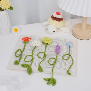 May include: Crocheted flower bookmarks in various colors, including red, white, yellow, green, and purple, are displayed on an open book. The handmade bookmarks feature detailed petals and green stems with leaves. A crocheted turtle and a vase with flowers are also in the image.