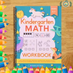 May include: A spiral-bound kindergarten maths workbook with a blue cover. The cover features the text "Kindergarten MATH WORKBOOK" with illustrations of a unicorn, sun, dinosaur, fox, and stars. The workbook is for ages 5-7.
