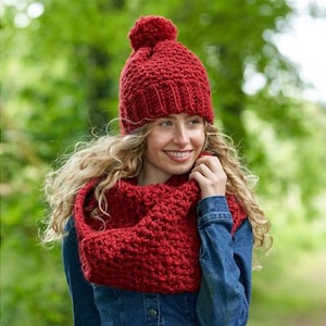 Puede incluir: Un gorro rojo tejido con pompón y un pañuelo circular a juego. El gorro y el pañuelo están hechos de un hilo grueso y texturizado. La mujer lleva una chaqueta vaquera.
