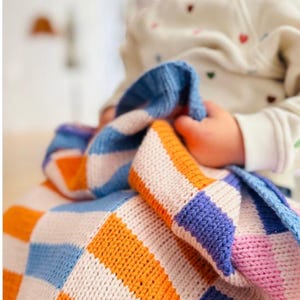 May include: A colorful, knitted blanket with a checkerboard pattern. The blanket features squares and stripes in shades of blue, orange, white, and pink. The blanket is soft and appears to be handmade. The blanket is being held by a person.