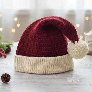 Puede incluir: Un gorro de Papá Noel burdeos y crema con un pompón blanco. El gorro es de punto y está sobre una superficie blanca, rodeado de piñas, vegetación y una taza de chocolate caliente. Decoración festiva navideña.
