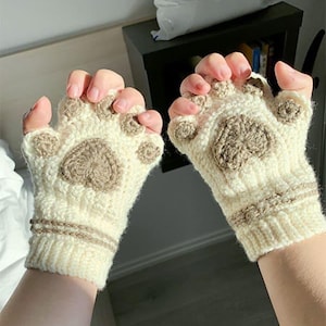 May include: A pair of cream-colored, fingerless gloves with a cat paw design. The gloves feature brown paw pads and a heart-shaped design on the palm. The wrist area has a brown knitted trim.