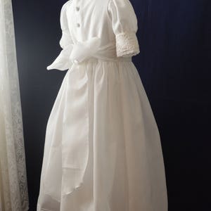May include: A white dress with a lace trim and a satin sash. The dress has a round neckline and short sleeves.