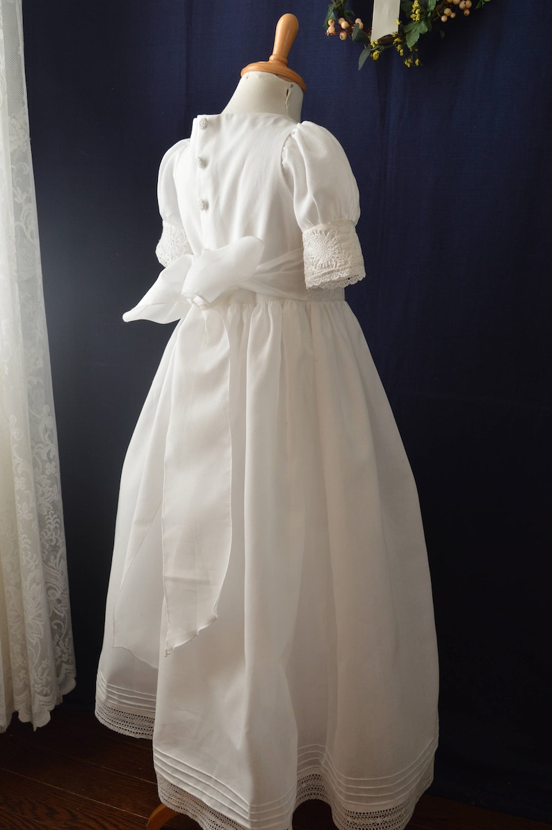 May include: A white dress with a lace trim and a satin sash. The dress has a round neckline and short sleeves.