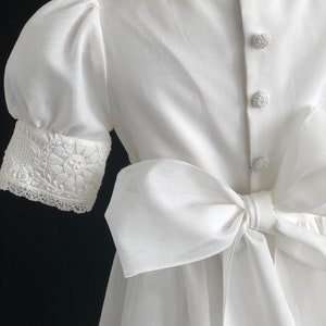 May include: White dress with a large bow in the back and lace trim on the sleeves.