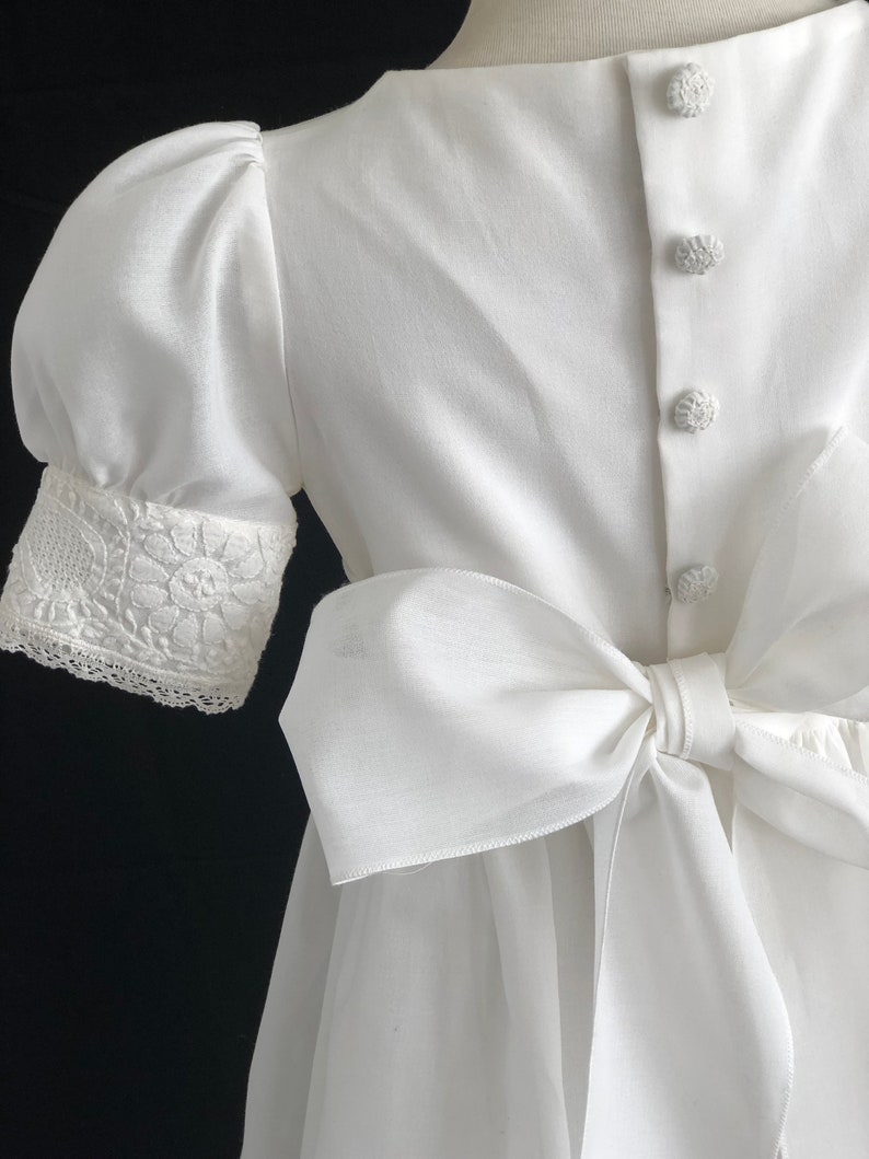 May include: White dress with a large bow in the back and lace trim on the sleeves.
