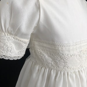 May include: A white cotton dress with lace trim and a gathered skirt. The dress has short puffed sleeves and a round neckline.