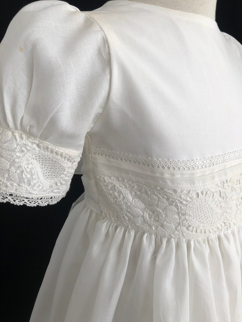 May include: A white cotton dress with lace trim and a gathered skirt. The dress has short puffed sleeves and a round neckline.