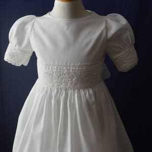 May include: A white dress with puffed sleeves and lace trim around the neckline and cuffs. The dress has a gathered skirt and a lace waistband.