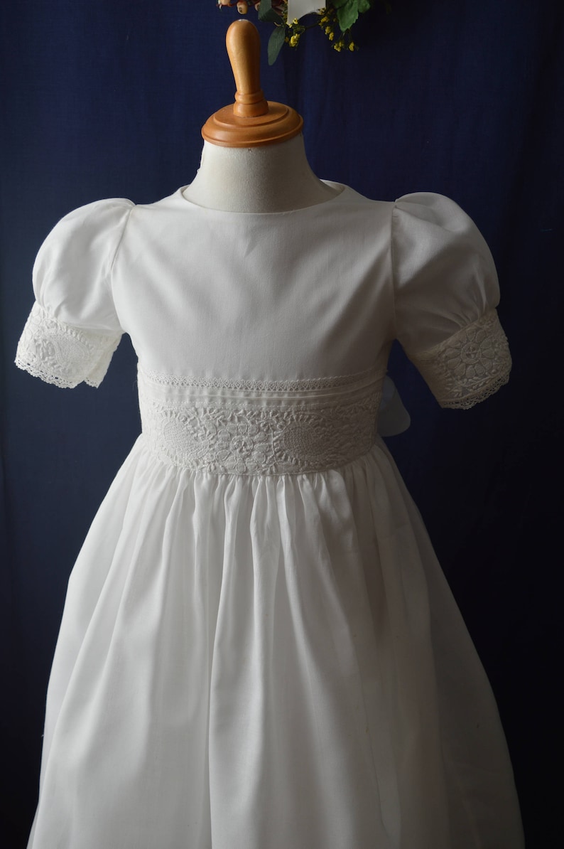 May include: A white dress with puffed sleeves and lace trim around the neckline and cuffs. The dress has a gathered skirt and a lace waistband.