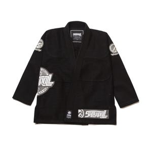 Pode incluir: Kimono de Jiu-Jitsu preto com logotipos brancos e cinzentos. O kimono apresenta gola de lapela, mangas compridas e costuras reforçadas. Os logotipos incluem a marca "SHOYOROLL" e outros elementos gráficos. O kimono está estendido sobre um fundo branco.