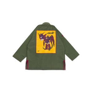 Op de afbeelding: Een olijfgroene jas met een grote rechthoekige gele patch op de achterkant. De patch toont een afbeelding van een paarse en rode robot met de tekst Mobile Suit Gundam W. De jas heeft lange mouwen en kleine rode zijsplitten aan de onderkant.