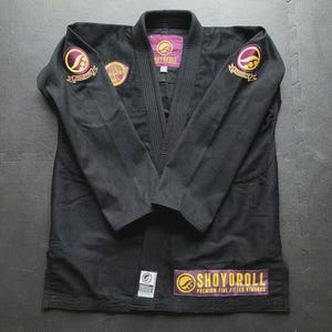 Puede incluir: Gi de Jiu-Jitsu negro con parches bordados morados y dorados. El gi presenta el nombre de la marca "SHOYOROLL" en la solapa y el dobladillo inferior. El gi está plano sobre una superficie gris.