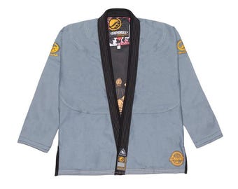 May include: A light blue martial arts gi with black trim and gold detailing. The gi displays the brand name "Shoyoroll" on the collar and patches. The interior lining has a floral pattern. This uniform is designed for martial arts training and competition.