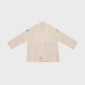 May include: Off-white martial arts gi with a V-neck design. The gi features embroidered patterns on the sleeves and lower front. The brand name is visible on the collar and lower front. The gi is laid flat against a light background.