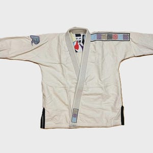 Könnte beinhalten: Cremefarbener Kampfsportanzug mit Kragen und langen Ärmeln. Die Uniform hat schwarze Akzente an den Manschetten und am Saum. Auf den Ärmeln sind Aufnäher mit geometrischen Designs und einem Logo zu sehen.