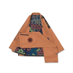 Op de afbeelding: Een beige vechtsport-gi met een donkerblauwe binnenvoering met een herhalend geometrisch bloemenpatroon. De buitenstof is effen beige met zwarte ronde patches en geborduurde details op de mouwen en de broekspijp.