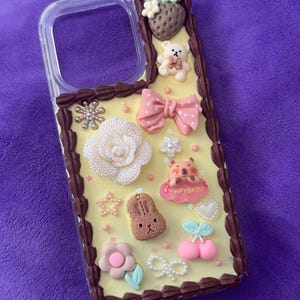 May include: A clear phone case decorated with various 3D embellishments. The design includes a strawberry, teddy bear, bow, flower, bunny, and a capybara figure. The case has a yellow background with a brown border.