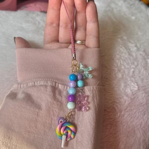 May include: A colorful phone charm with a pink string, rainbow lollipop, purple and blue beads, gummy bear charms, and a light blue butterfly charm.