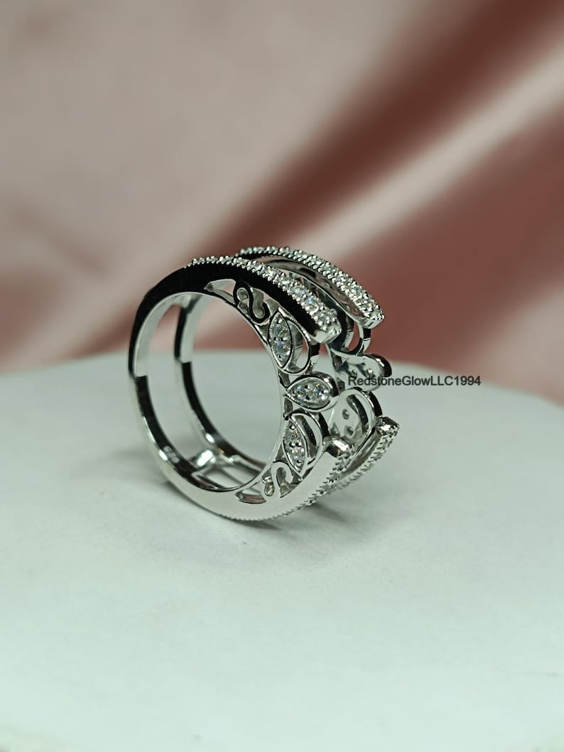 Puede incluir: Un anillo de plata con un dise&ntilde;o delicado e intrincado con peque&ntilde;as piedras transparentes. El anillo tiene una banda doble y est&aacute; sobre un fondo blanco.