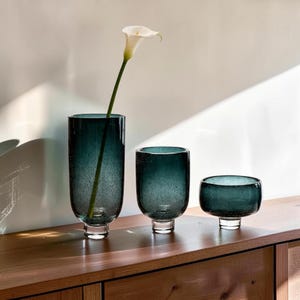 May include: Three cylindrical glass vases in varying heights, featuring a deep teal blue color with a subtle bubble texture throughout the glass. Each vase has a clear, thick circular base. A single white calla lily is placed in the tallest vase.