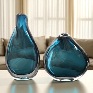Könnte beinhalten: Zwei tropfenförmige Glasvasen in Teal-Farbe. Die größere Vase ist schlank und hoch, die kleinere rundlicher. Beide haben eine glänzende Oberfläche und stehen auf einer weißen Fläche. Höhe der großen Vase: ca. 30 cm.