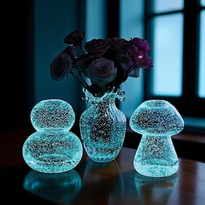 Pode incluir: Natureza morta com um vaso de vidro com um buquê de rosas escuras, ladeado por dois objetos decorativos de vidro. Os objetos são uma esfera e um formato de cogumelo, todos com um brilho azul e textura de bolhas. A cena é ambientada em uma superfície de madeira escura.