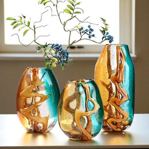 Op de afbeelding: Drie decoratieve glazen vazen in tinten van turquoise en amber, met een organisch, vloeiend ontwerp. De vazen hebben verschillende maten en vormen, met een takje met kunstmatige bessen en bladeren in één. Een stijlvol woonaccessoire.
