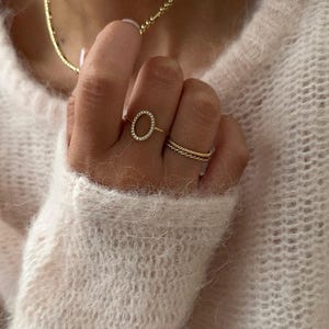 Peut inclure: Une bague en or avec un cercle de petits diamants autour d'une forme ovale. La bague est portée à l'index d'une main portant un collier en chaîne dorée.