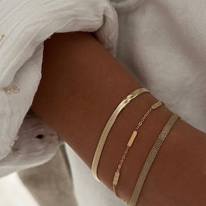 Puede incluir: Tres pulseras de oro apiladas en una muñeca. La pulsera superior es una cadena de serpiente gruesa y plana. La pulsera del medio es una cadena delicada con pequeños abalorios rectangulares. La pulsera inferior es una cadena de malla fina.