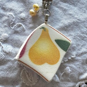 Könnte beinhalten: Eine silberne Halskette mit einem Anhänger aus einem Stück Vintage-Keramik. Der Anhänger zeigt ein gelbes Birnen-Design mit grünen und roten Akzenten. Zwei kleine gelbe Perlen hängen an der Halskette.