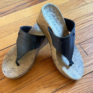 以下が含まれることがあります： コルクスタイルのウェッジヒールが付いた、黒い革製のトングサンダルのペア。サンダルはトングストラップのデザインとテクスチャード加工されたフットベッドを備えています。ウェッジヒールはサポートを提供します。サンダルは木製の表面に置かれています。