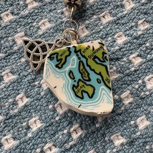 May include: A silver-toned necklace with a pendant featuring a map design. The pendant is a white, irregular shape with a blue and green map design. A silver Celtic knot charm hangs next to the pendant. The necklace is on a blue and white patterned surface.