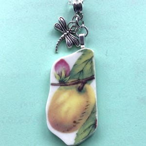 May include: A silver chain necklace with a pendant featuring a yellow pear with a pink blossom and green leaves. A silver dragonfly charm hangs from the top of the pendant.