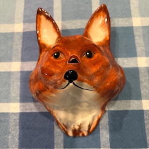Könnte beinhalten: Ein Wandobjekt aus Keramik in Form eines Fuchskopfes mit glänzender Oberfläche in Orange-, Braun- und Weißtönen. Der Fuchs hat spitze Ohren, dunkle Augen, eine schwarze Nase und ein weißes Kinn. Die Skulptur ist zur Wandmontage gedacht.