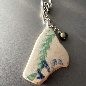 May include: A unique necklace featuring a piece of sea pottery with green and blue designs. The pendant is attached to a silver chain and accented with a small, dark pearl. The pottery has a cream-colored base.