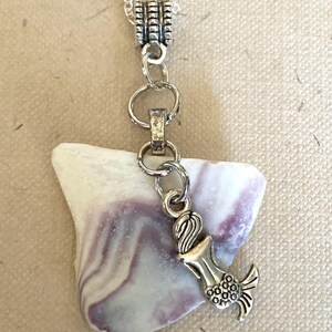 Peut inclure: Un collier argenté avec un pendentif en verre de mer blanc et violet. Un petit charme de sirène argenté est suspendu en dessous de la chaîne. Le collier est présenté sur une surface texturée blanc cassé.