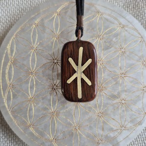 May include: A dark brown wooden pendant with a gold rune symbol, suspended from a black cord. The pendant is rectangular with rounded corners. It hangs above a white, circular surface with a gold floral design.