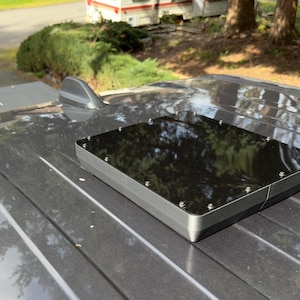 Puede incluir: Una caja rectangular negra con un acabado brillante está montada en el techo de un coche. La caja tiene varios tornillos que la sujetan al techo.