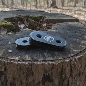 May include: Two black pulleys with a gear logo and text are stacked on a tree stump. The pulleys have two holes and a curved design. The background is a blurred forest scene with sunlight.